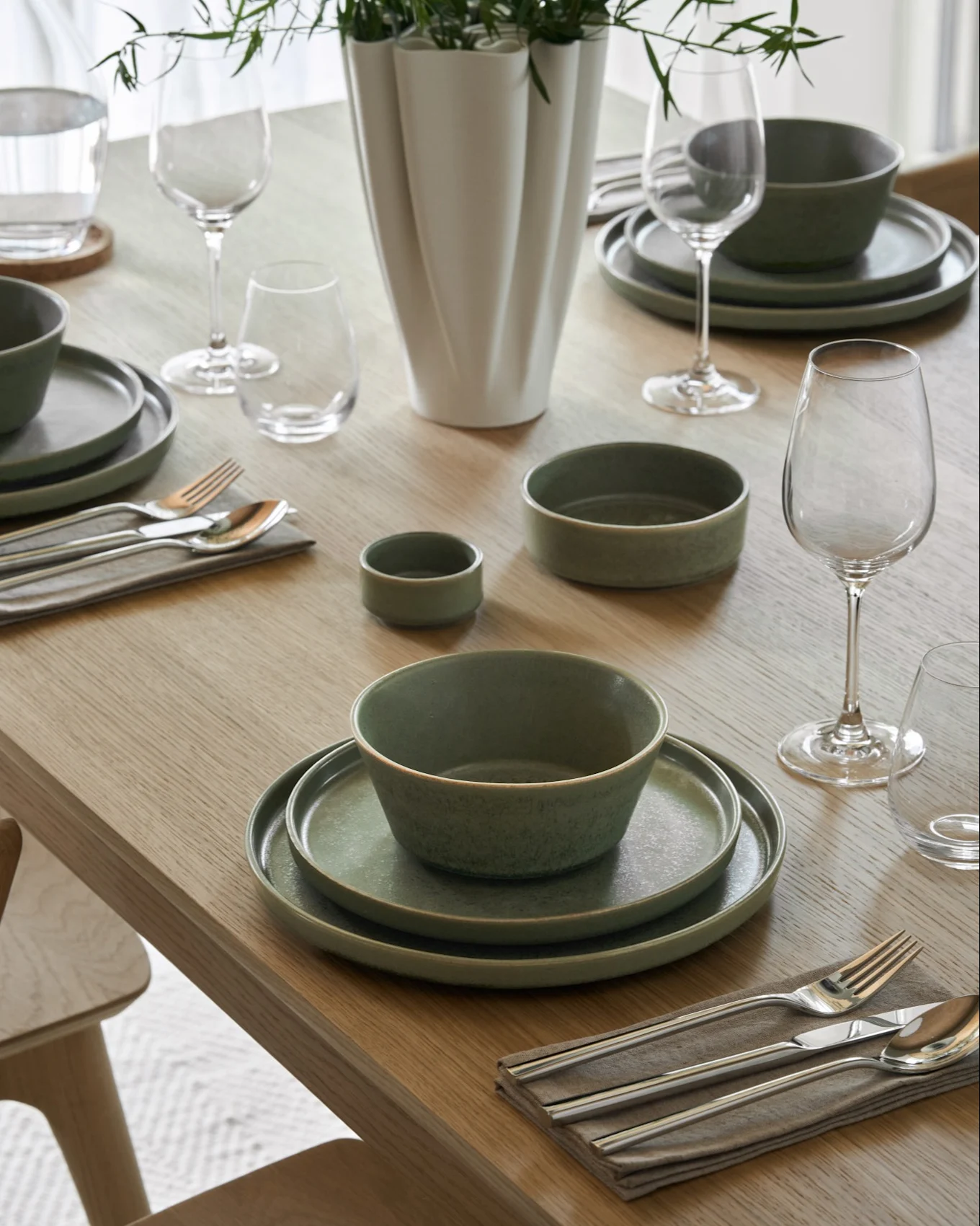 Wooden dining table set with bowls of creamy soup, matching plates, wine and water glasses, silverware, and a vase with green foliage.