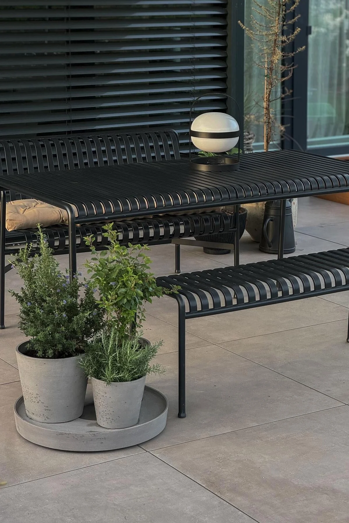 Black metal outdoor dining set with slatted table and benches, spherical lantern, and potted green plants on a patio.
