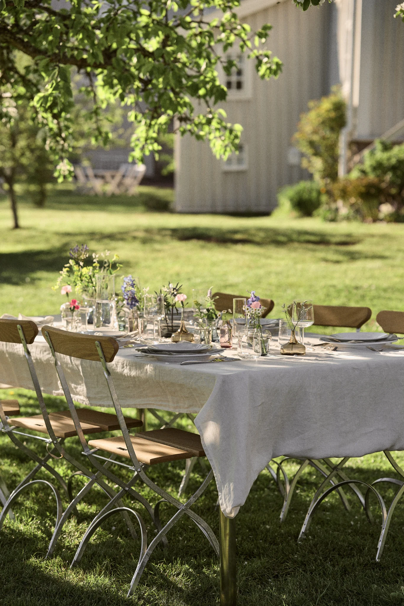 An outdoor dining table set with white plates, glasses, and floral arrangements, surrounded by folding chairs on a sunny lawn.
