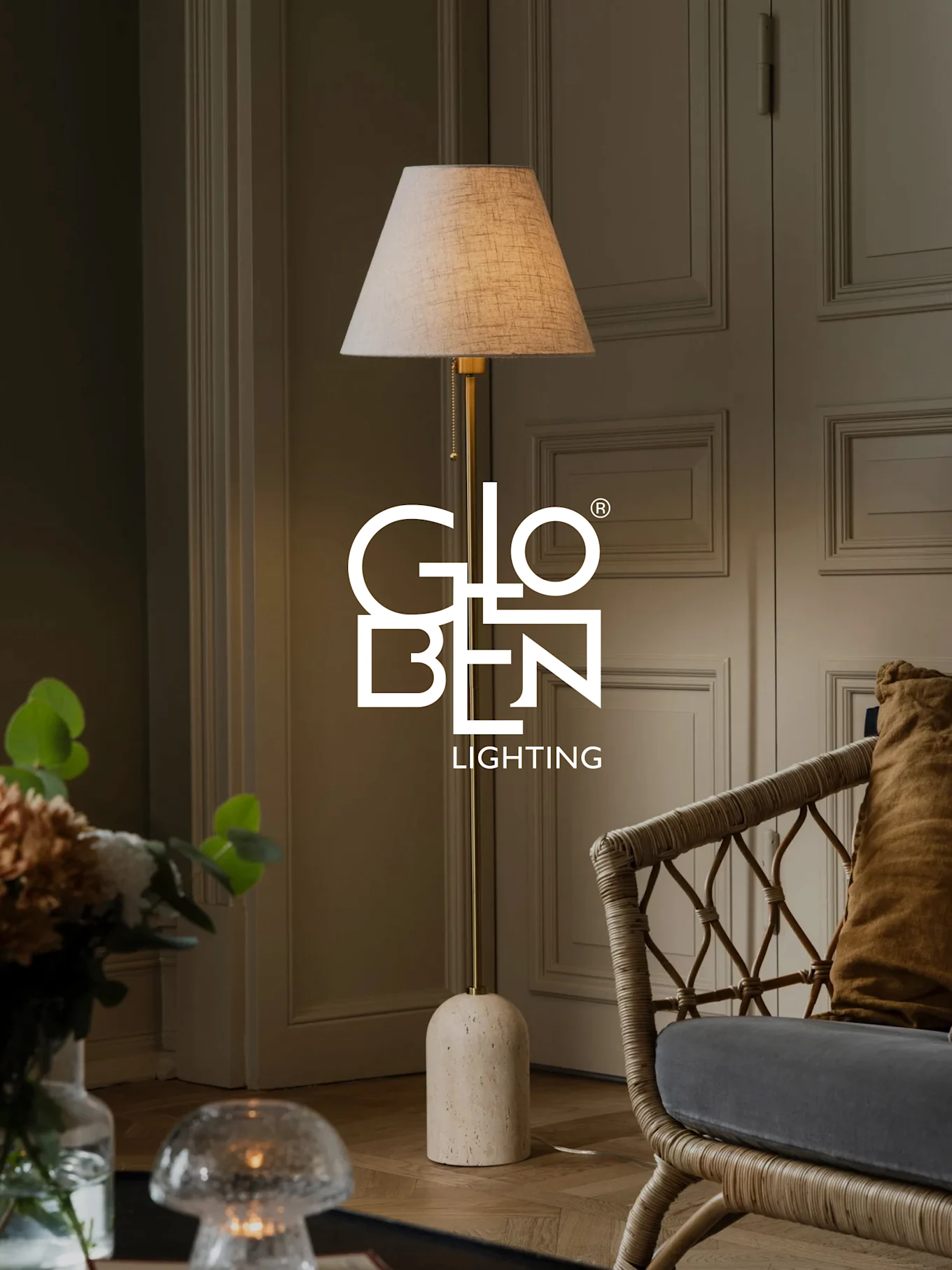 A floor lamp with a beige shade, brass stand, and rounded stone base stands next to a wicker chair.