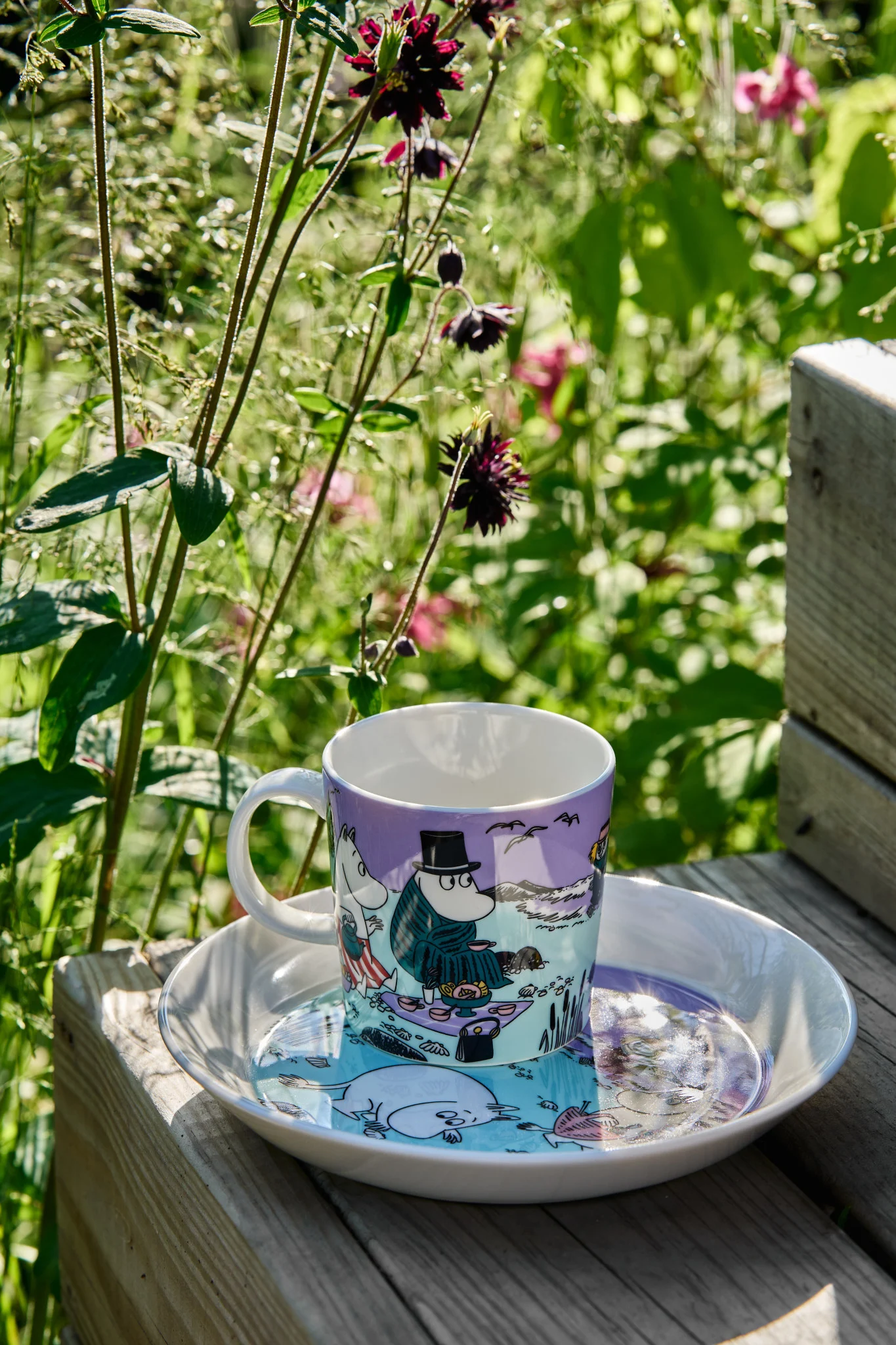 The summer Moomin mug 2025 ‘Beach Day’ stands on a step with the matching plate, with a wild meadow in the background.