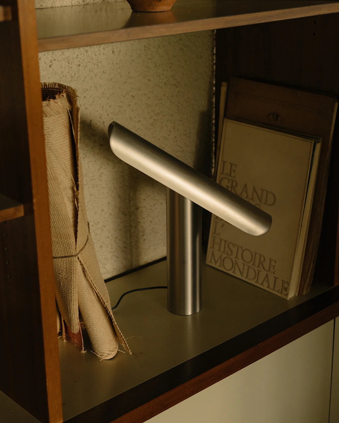 Modern silver desk lamp with a cylindrical base and an angled horizontal light bar on a shelf.