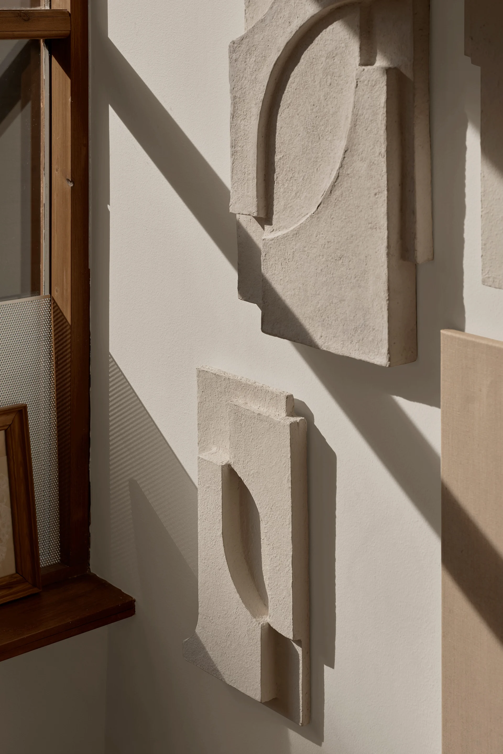 Textured, light-colored abstract wall art pieces with geometric shapes on a white wall, cast in strong diagonal sunlight.