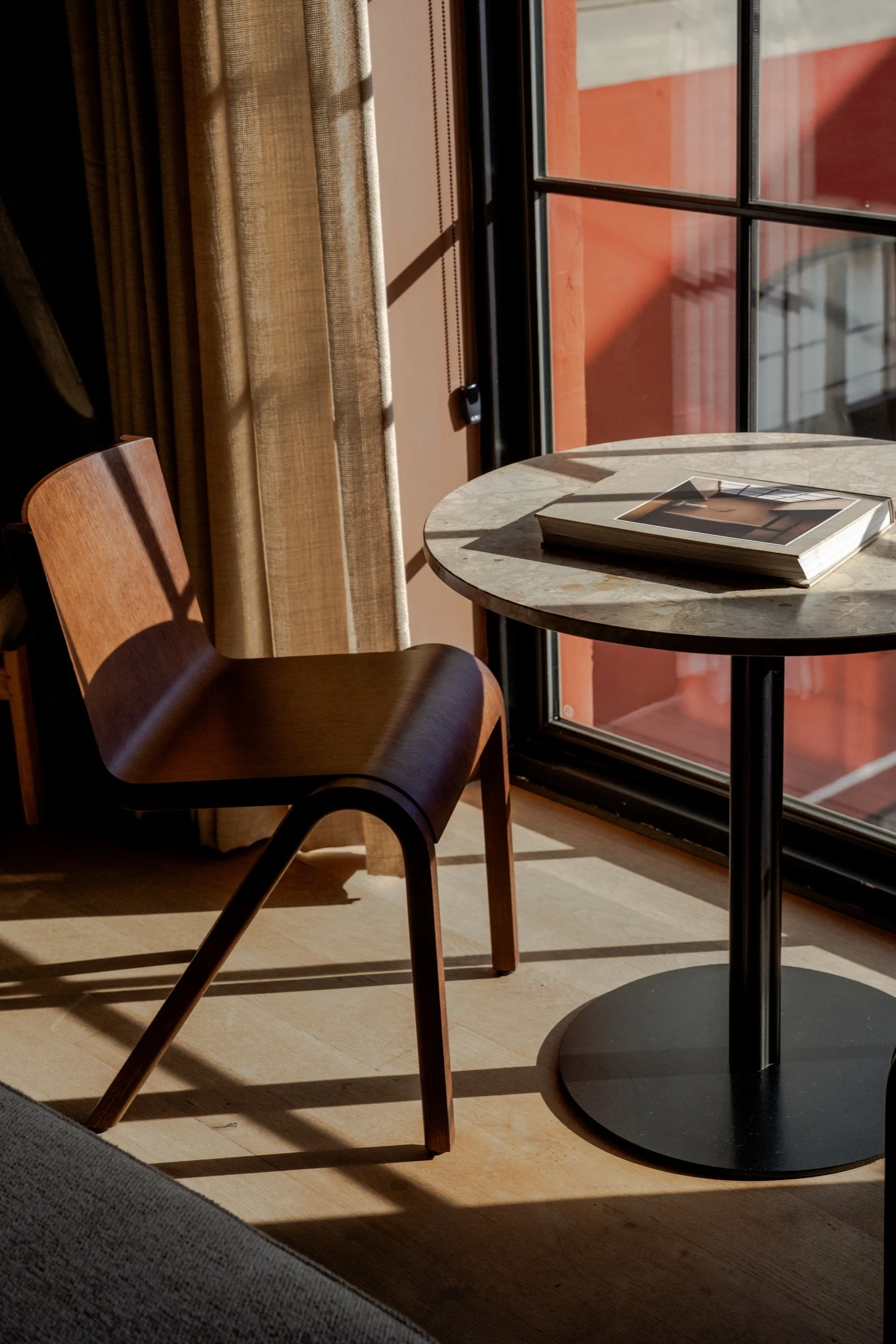 Modern wooden chair and round table with a book, bathed in sunlight by a window.