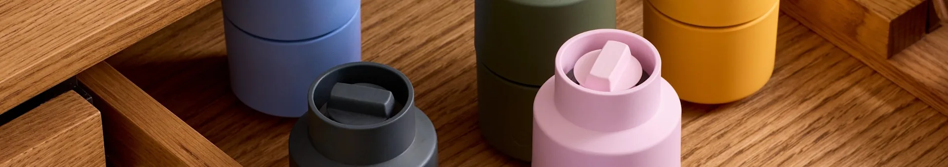 Five colorful containers: dark grey, light blue, olive, light pink with a unique cap, and mustard yellow, on wood.