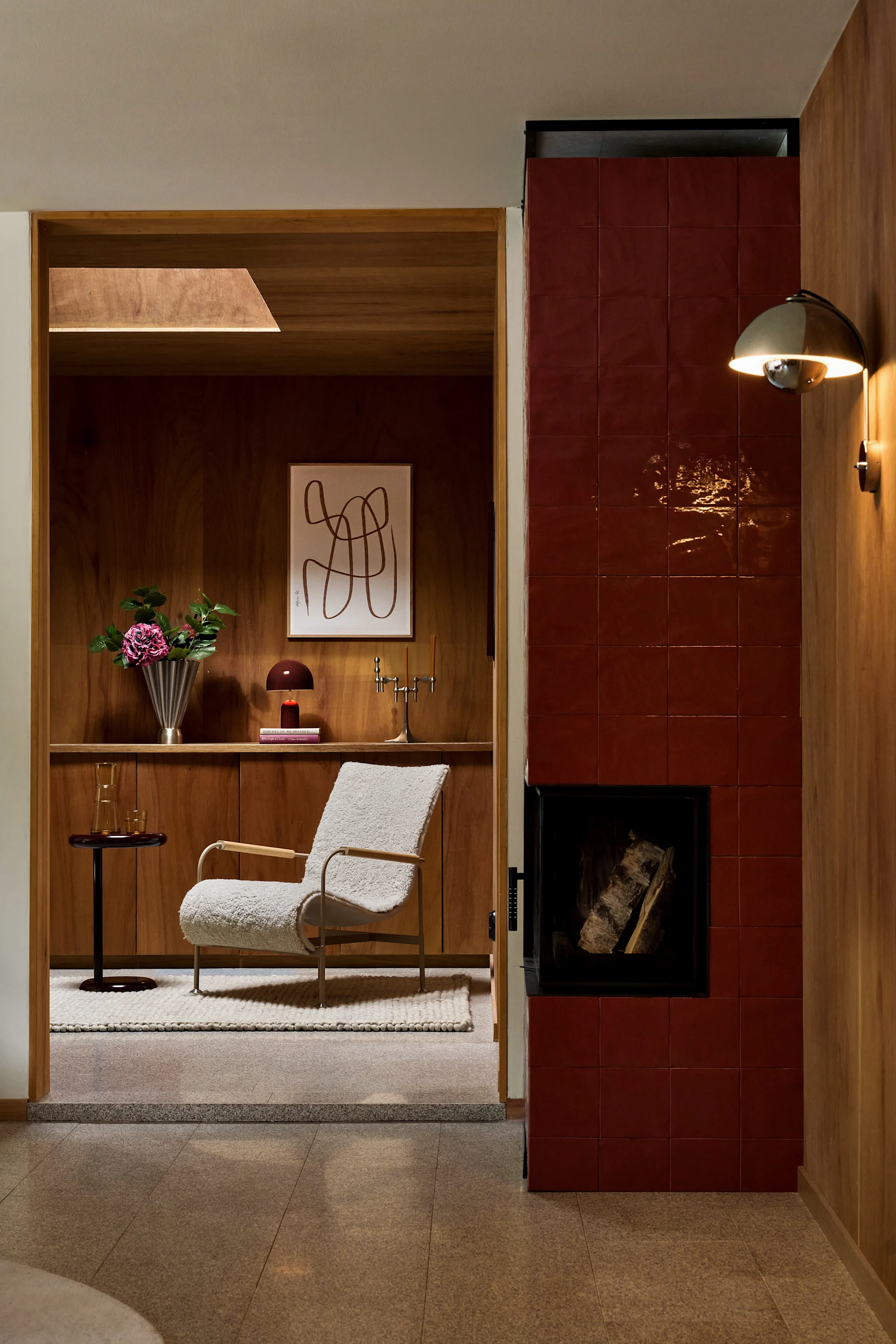 A cozy living area featuring a red tiled fireplace, a cream bouclé armchair, and wooden wall paneling with built-in shelves.