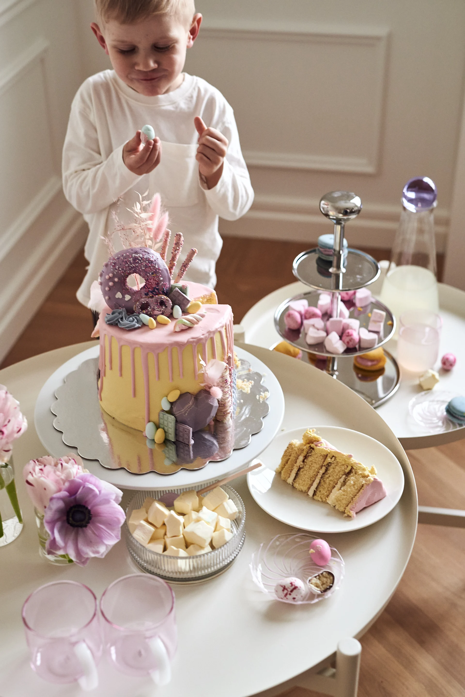 A boy holds a small blue candy in front of a table with a pink and yellow drip cake, marshmallows, and other desserts.
