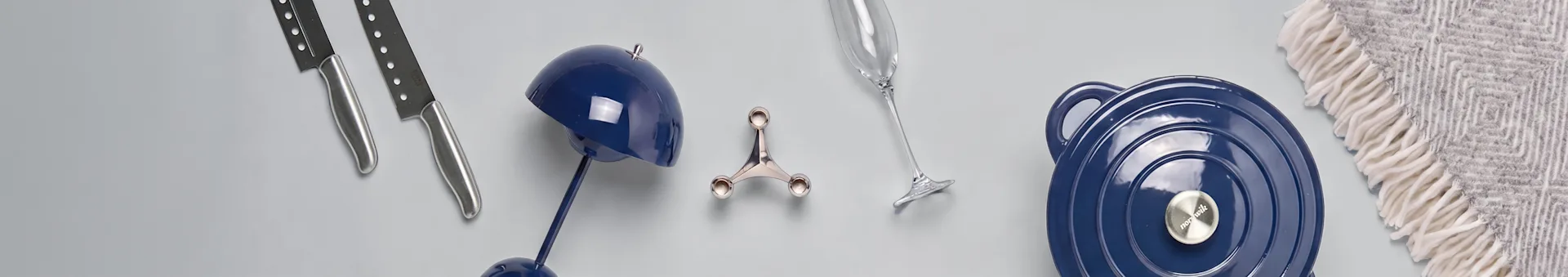 Two knives, a blue lamp, a silver holder, a champagne flute, a blue pot, and a grey patterned blanket on a light surface.