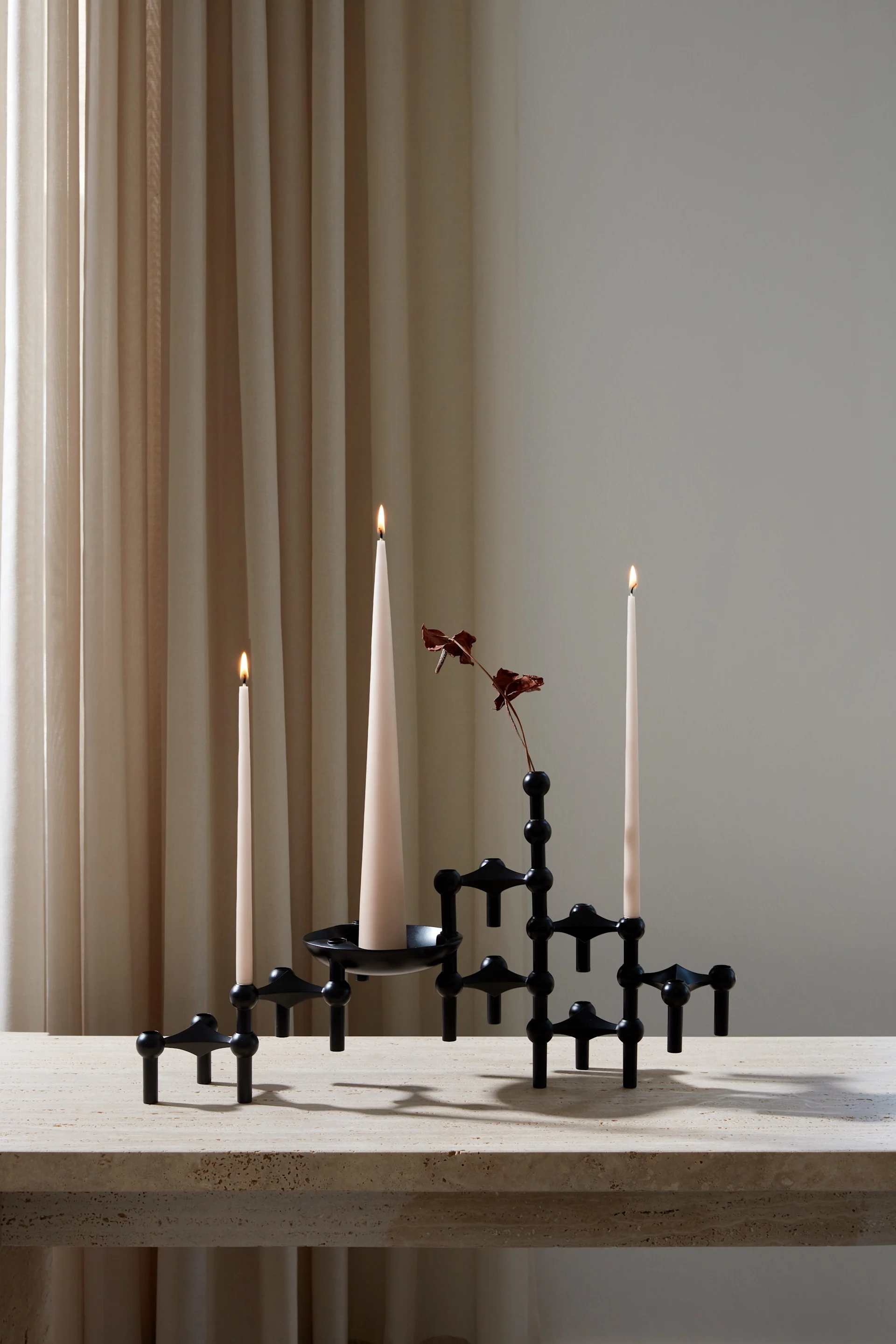 Black modular candelabra with three lit cream taper candles and a dried red branch on a beige stone table, with light curtains.
