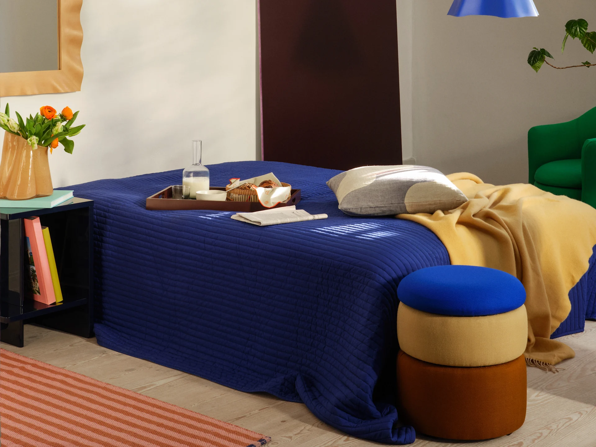 Modern bedroom with a blue quilted bedspread, yellow throw, breakfast tray, and stacked colorful ottomans.