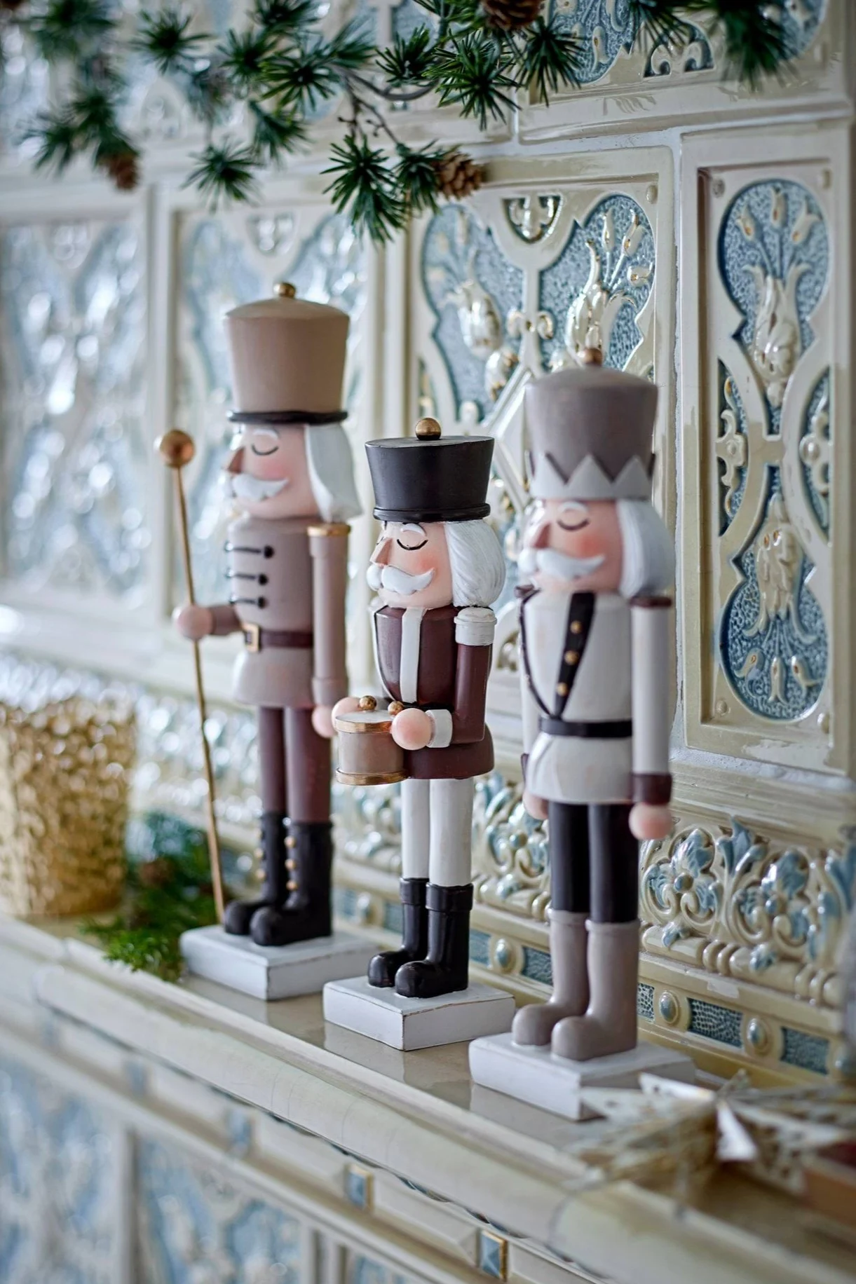 Three decorative nutcracker figures in varying brown, white, and grey uniforms stand on an ornate mantelpiece, adorned with pine branches and pinecones.