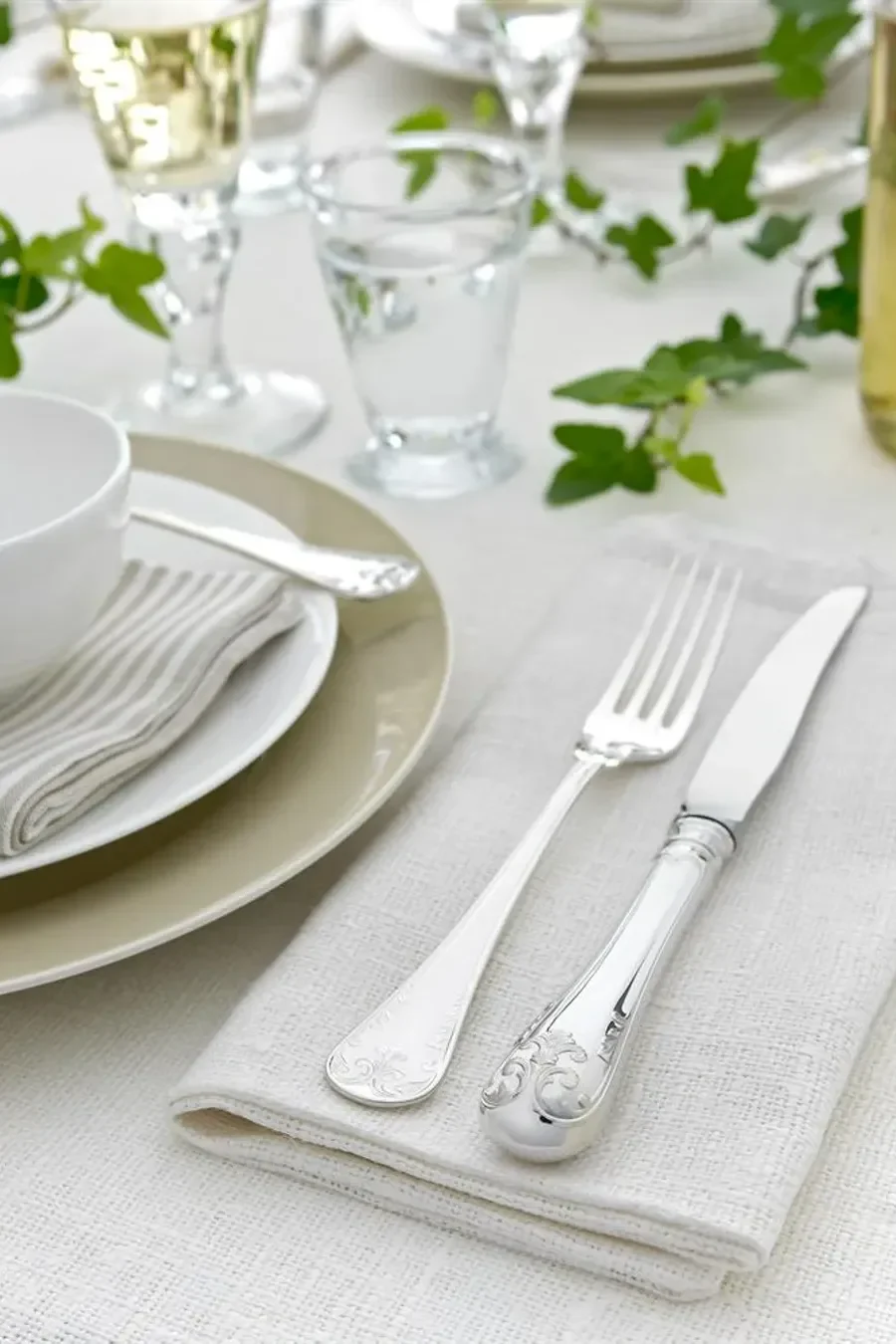 Elegant table setting with ornate silver cutlery, stacked plates, and glasses, adorned with green ivy on a white tablecloth.