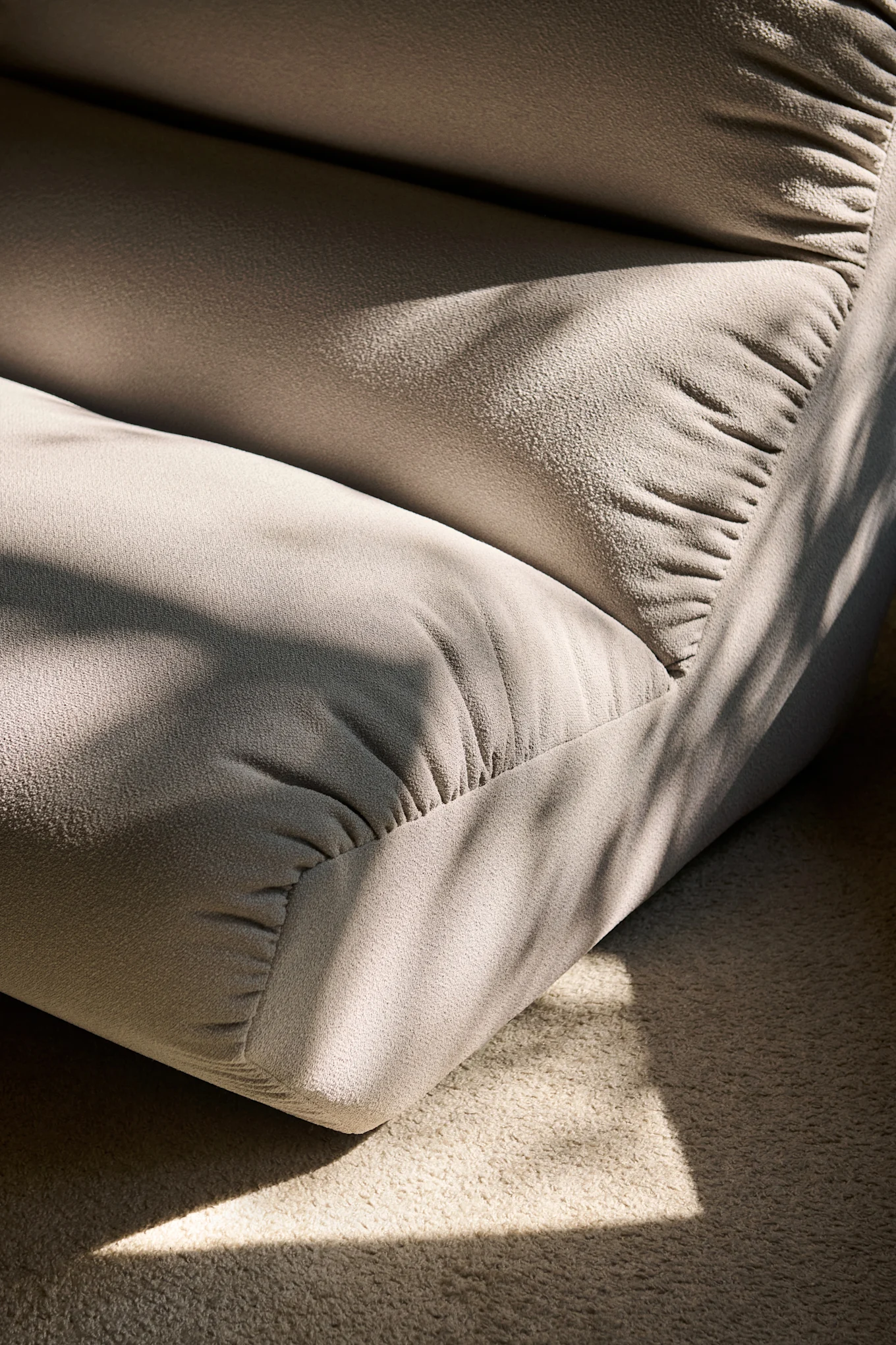 Light beige upholstered seating with deep folds and a textured surface, bathed in sunlight with patterned shadows on it and the carpet.