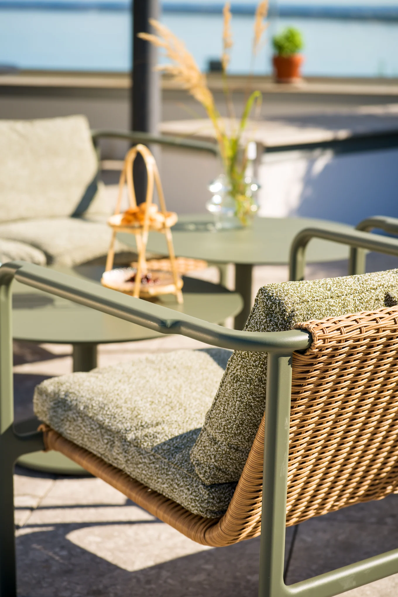 Outdoor patio chair with olive green frame, woven rattan back, and green cushions. Matching table and chair in blurry background near water.