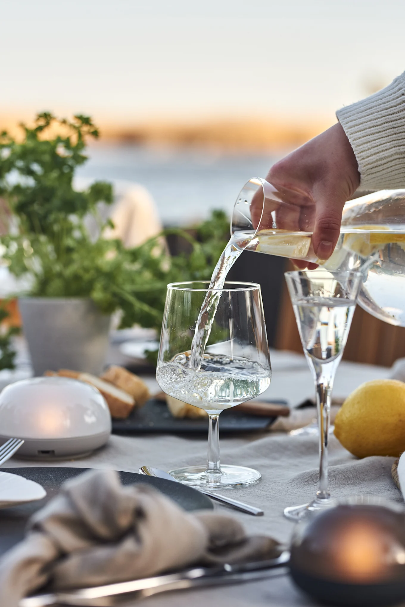 A hand in a white sweater pours water with lemon slices from a glass pitcher into a wine glass on an outdoor dining table.