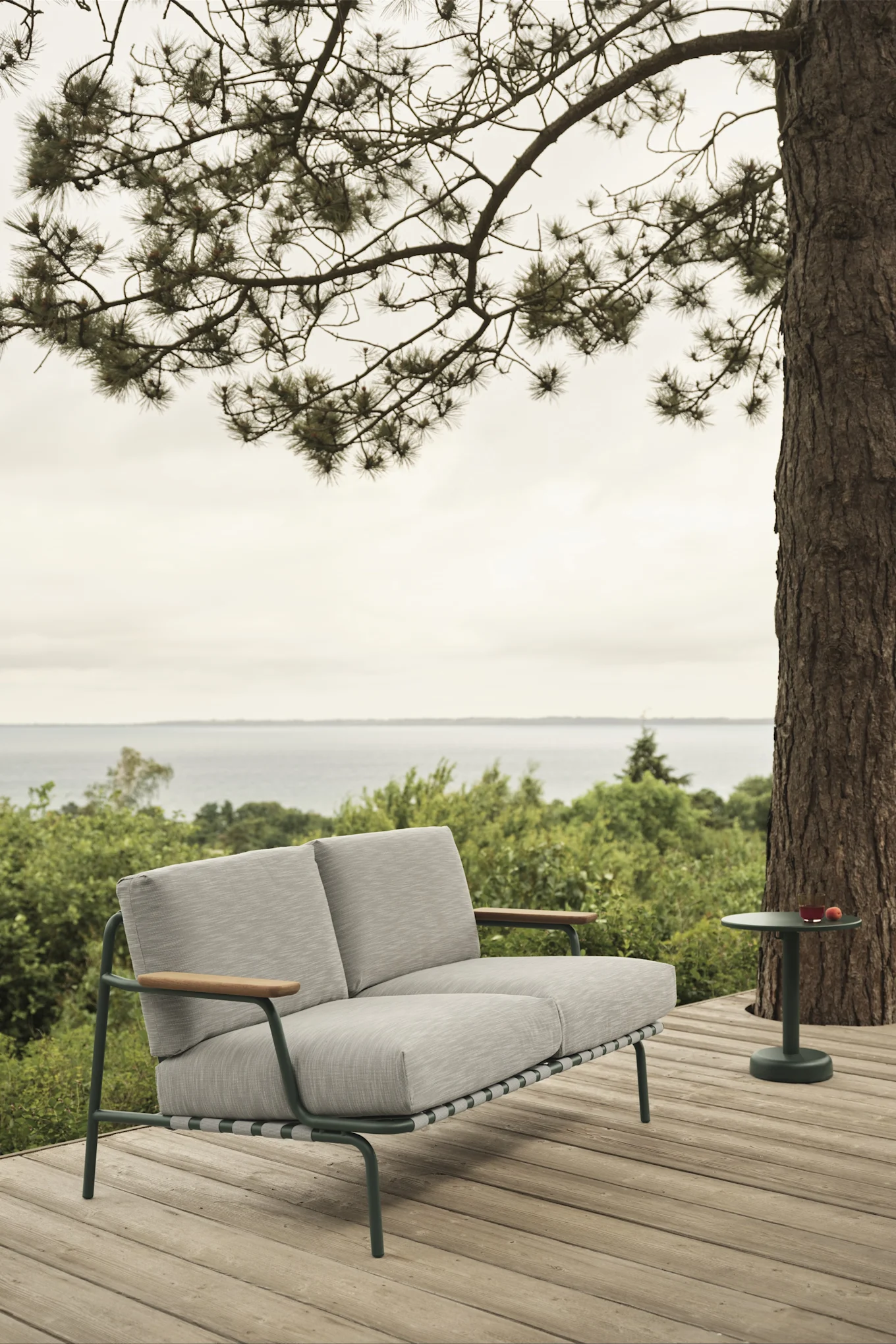 Outdoor scene with a light grey two-seater sofa on a wooden deck, a green side table with a drink, and a sea view.