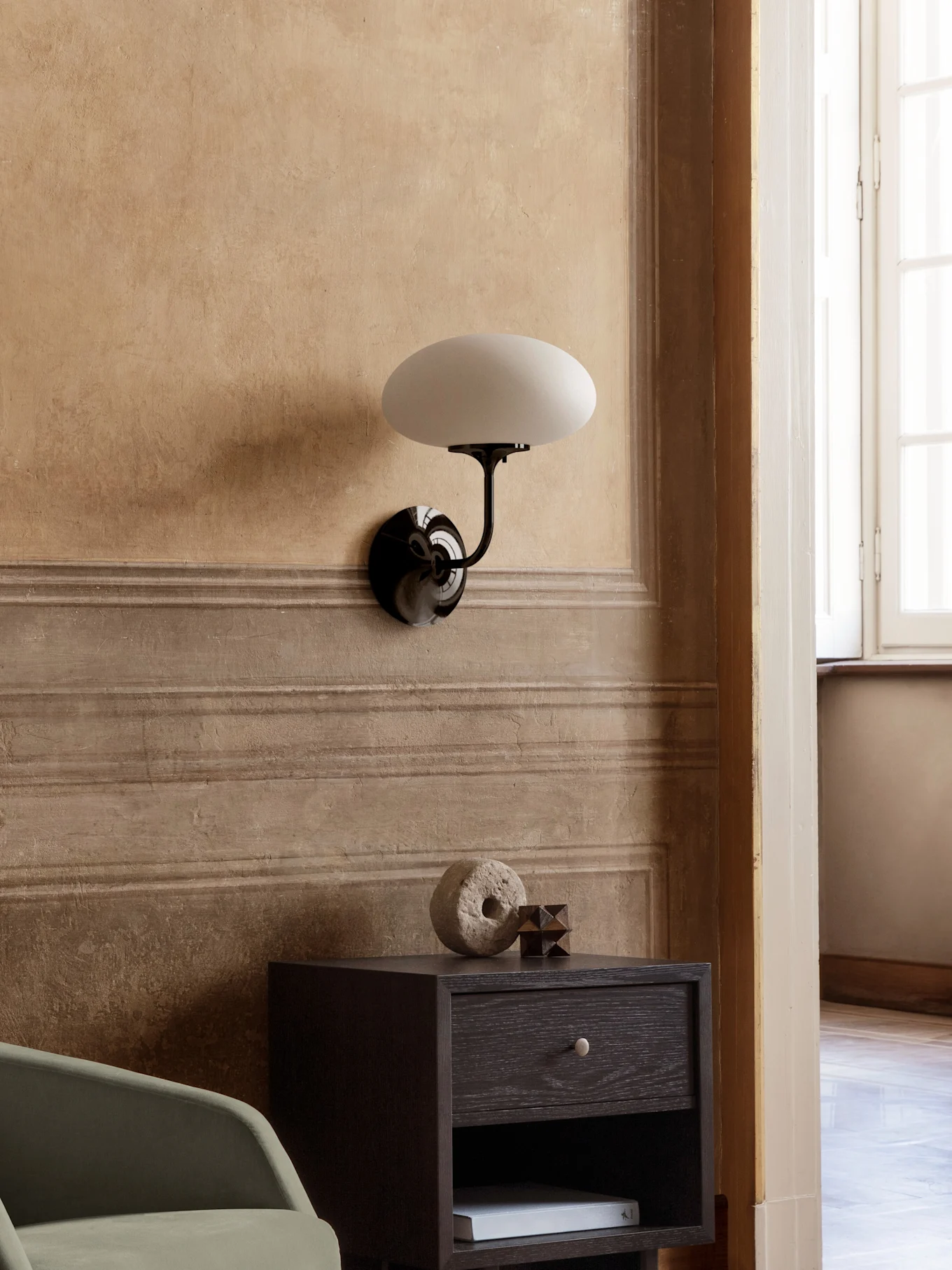 A wall lamp with a black arm and oval white shade above a dark wood nightstand with stone and wood decor.