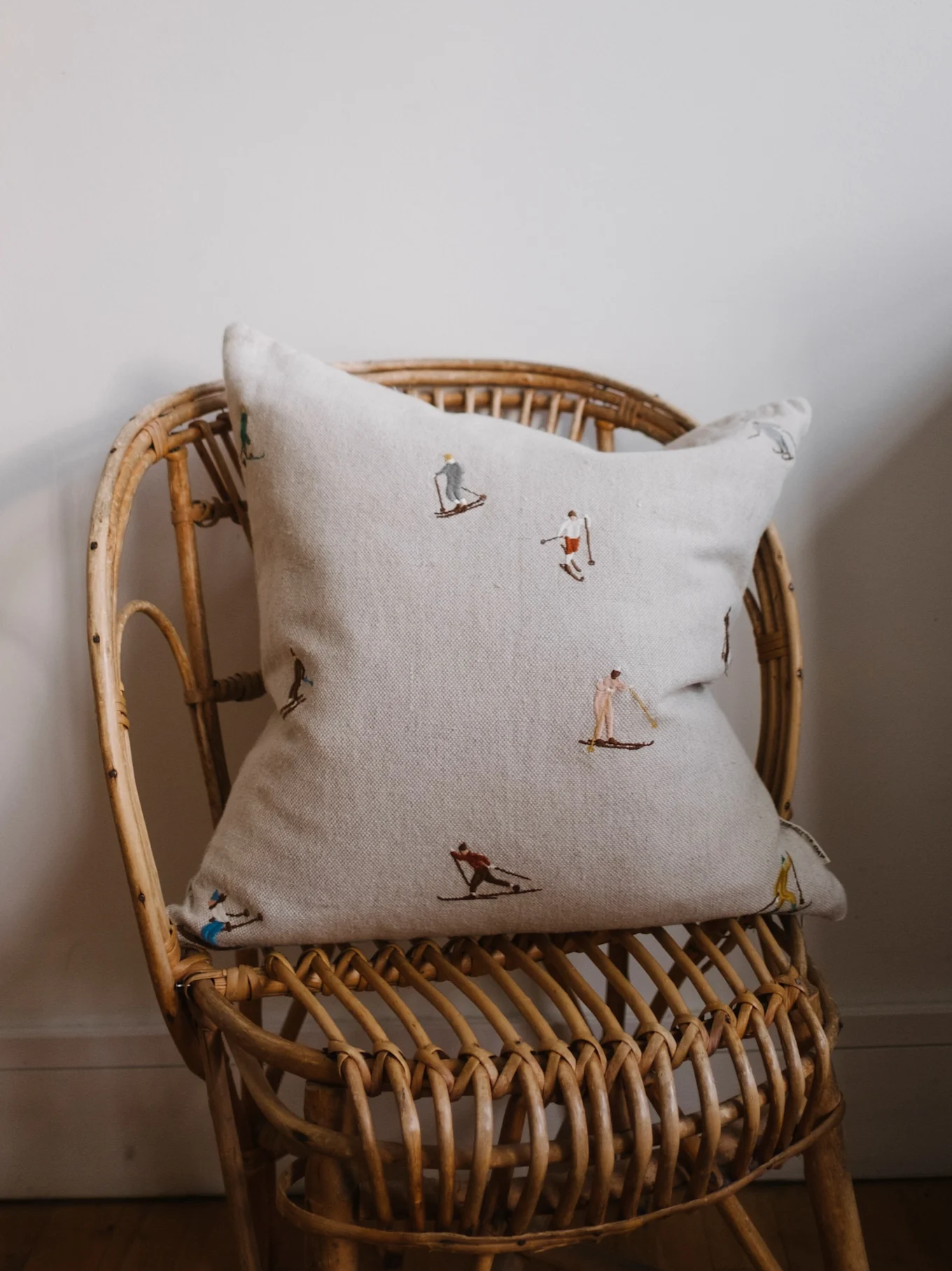 Light beige cushion with embroidered skiing figures on a wicker chair against an off-white wall.