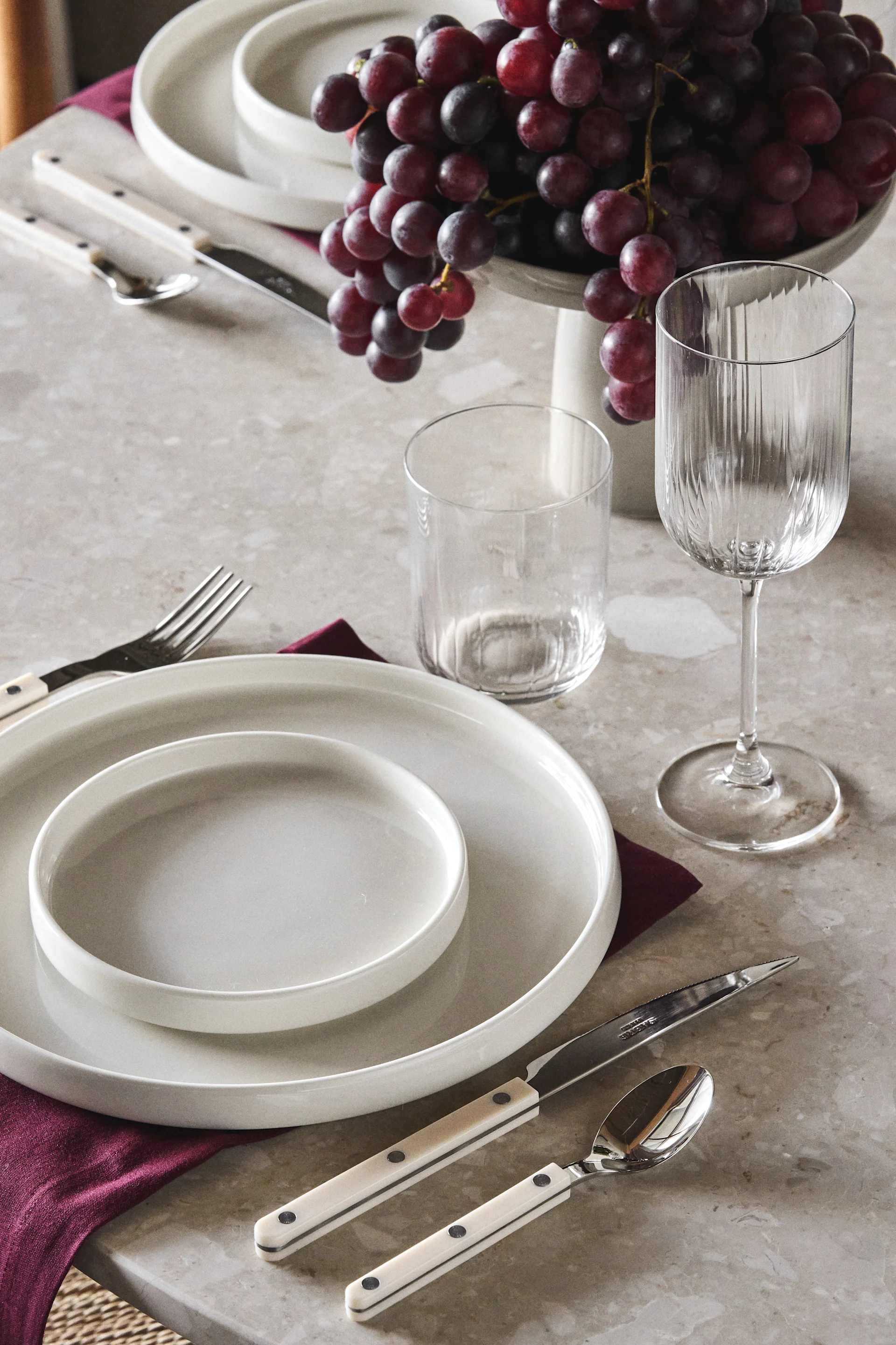 Elegant table setting with white plates, white-handled cutlery, two glasses, and a bowl of red grapes on a stone-colored table.