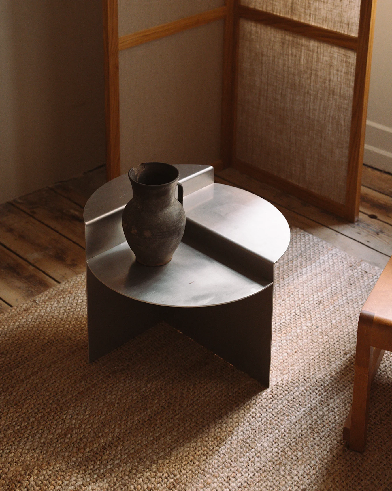 A minimalist, low, round silver metal coffee table with a rustic dark ceramic vase on a woven rug.