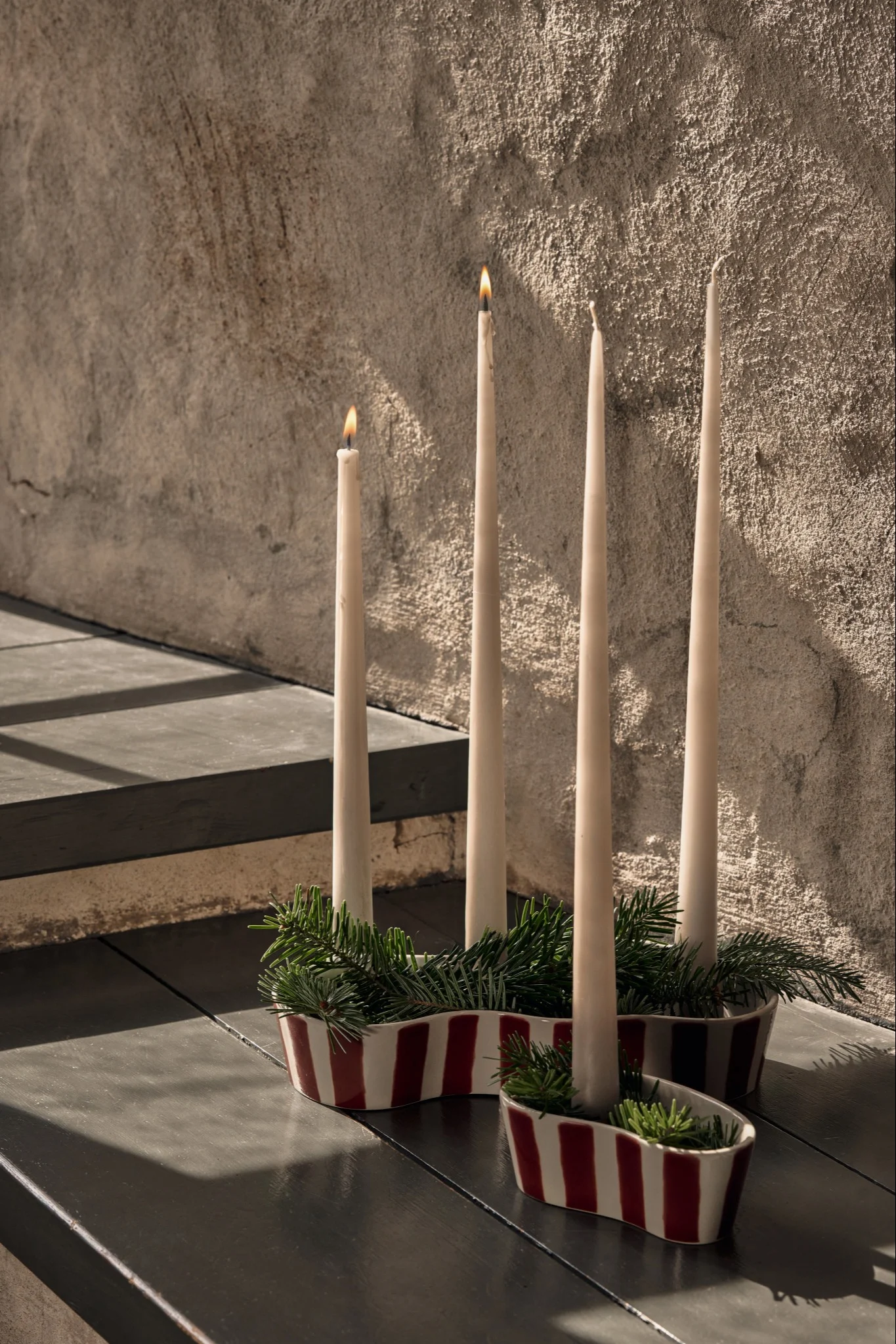 An advent wreath with two lit and two unlit white taper candles in a red and white striped, wave-shaped holder, accented with pine branches.