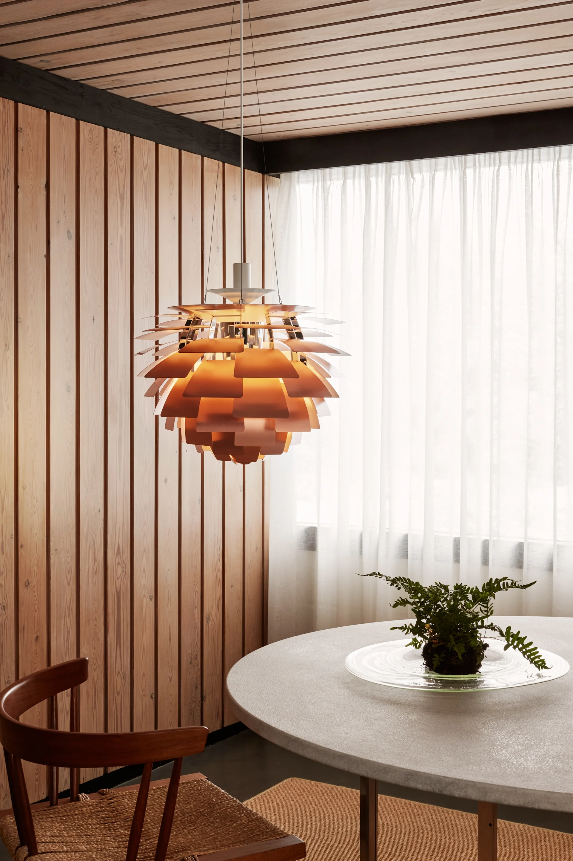 A copper layered pendant lamp hangs above a round grey table with a fern, next to wooden walls and sheer white curtains.