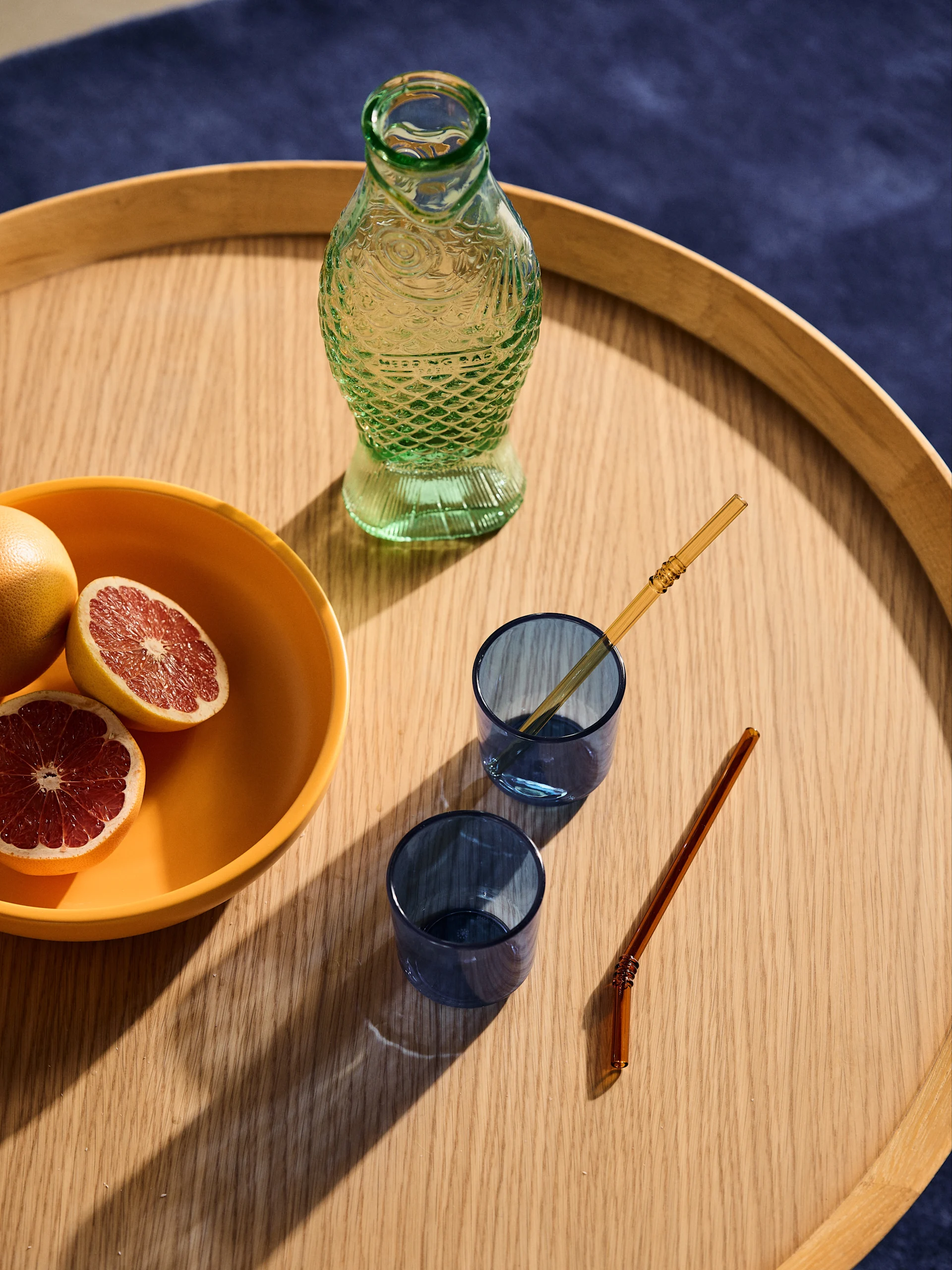 A wooden tray holds a green patterned glass bottle, an orange bowl with grapefruits, two blue glasses, and two gold straws.