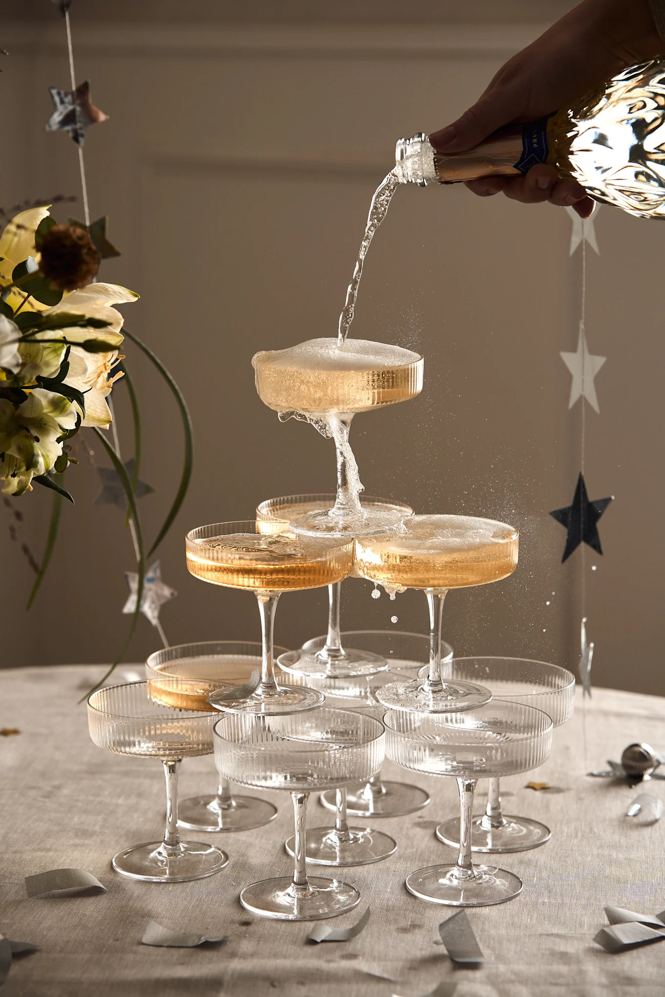A hand pours champagne into a tower of ribbed coupe glasses, overflowing with bubbly liquid onto a table with star decorations.