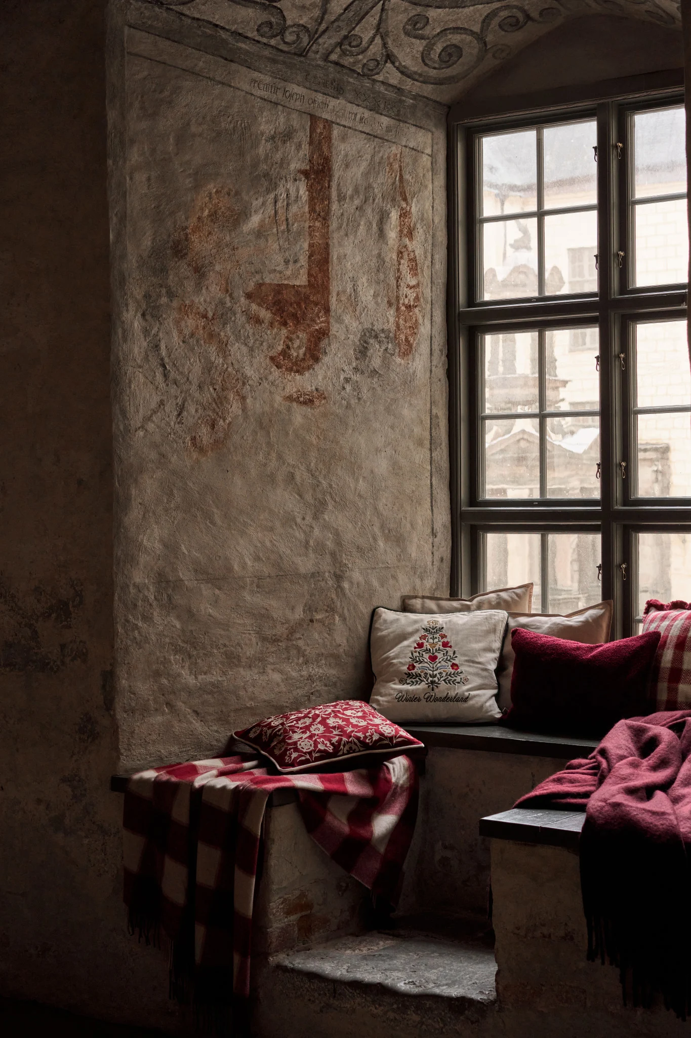 A cozy window nook in a rustic room, with embroidered and plaid cushions, and warm blankets on stone benches.