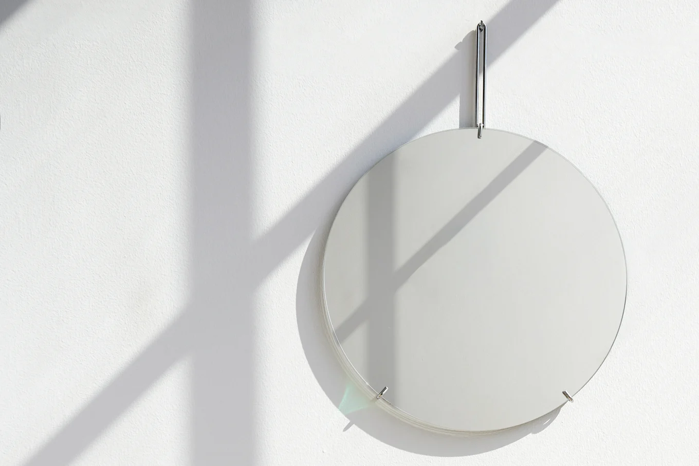 A round, clear glass panel hangs on a white textured wall, with window shadows falling across it and producing a subtle rainbow refraction.