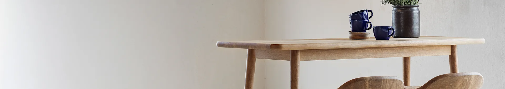 Light wooden table with blue mugs on coasters, a dark vase with herbs, and partial views of wooden chairs.