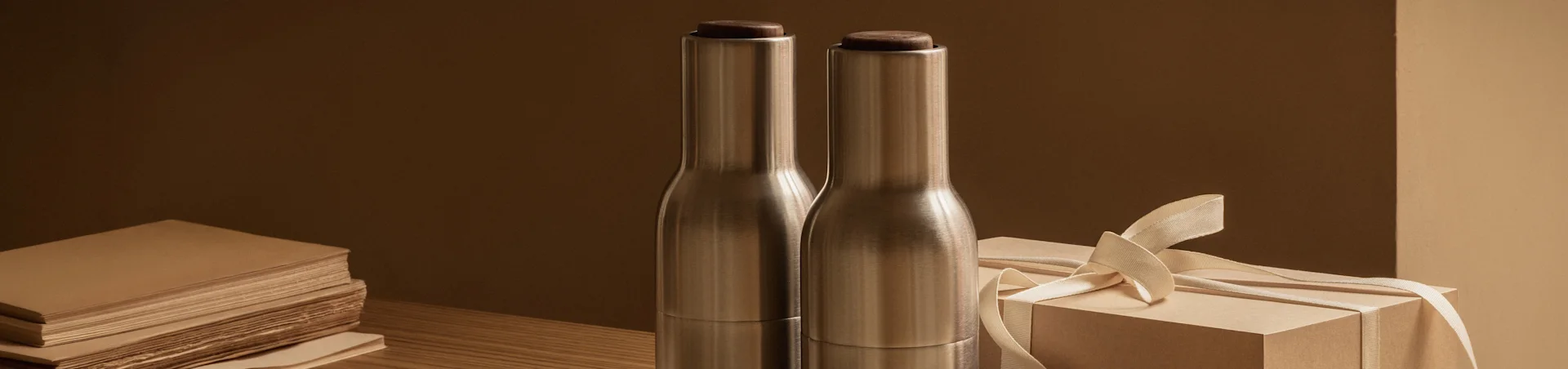Two brushed metal bottles with dark wooden tops next to a light brown gift box with a ribbon on a wooden table.