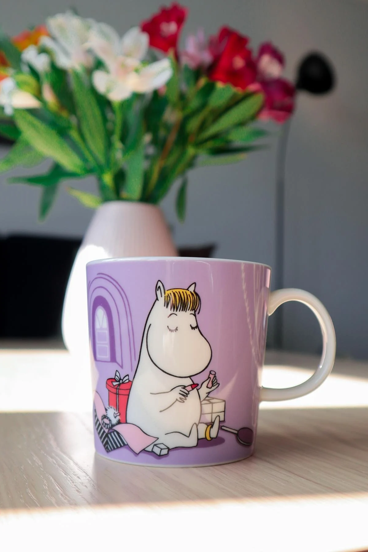 The purple Moomin mug with the Snorkmaiden on it stands on a table, in the background is a vase with a bunch of flowers.
