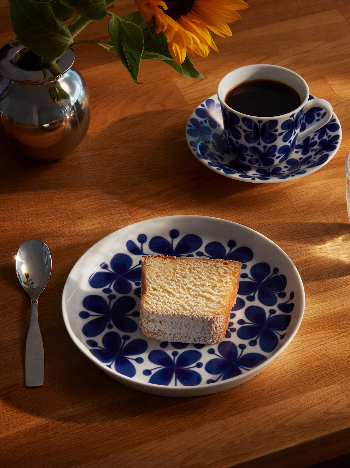 A slice of cake on a blue and white floral plate, with a coffee cup, glass of water, spoon, and a sunflower in a silver vase on a sunlit wooden table.