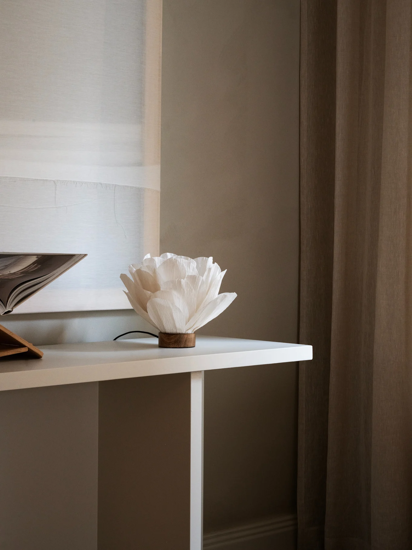 White flower-shaped paper lamp with a wooden base on a white table, next to an open book. Brown curtains in background.