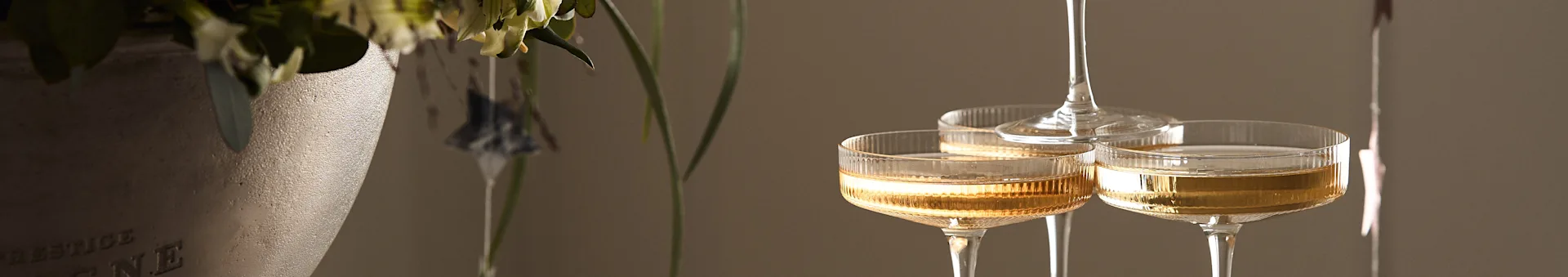 Three ribbed champagne coupe glasses filled with golden sparkling wine, beside a textured vase with flowers.