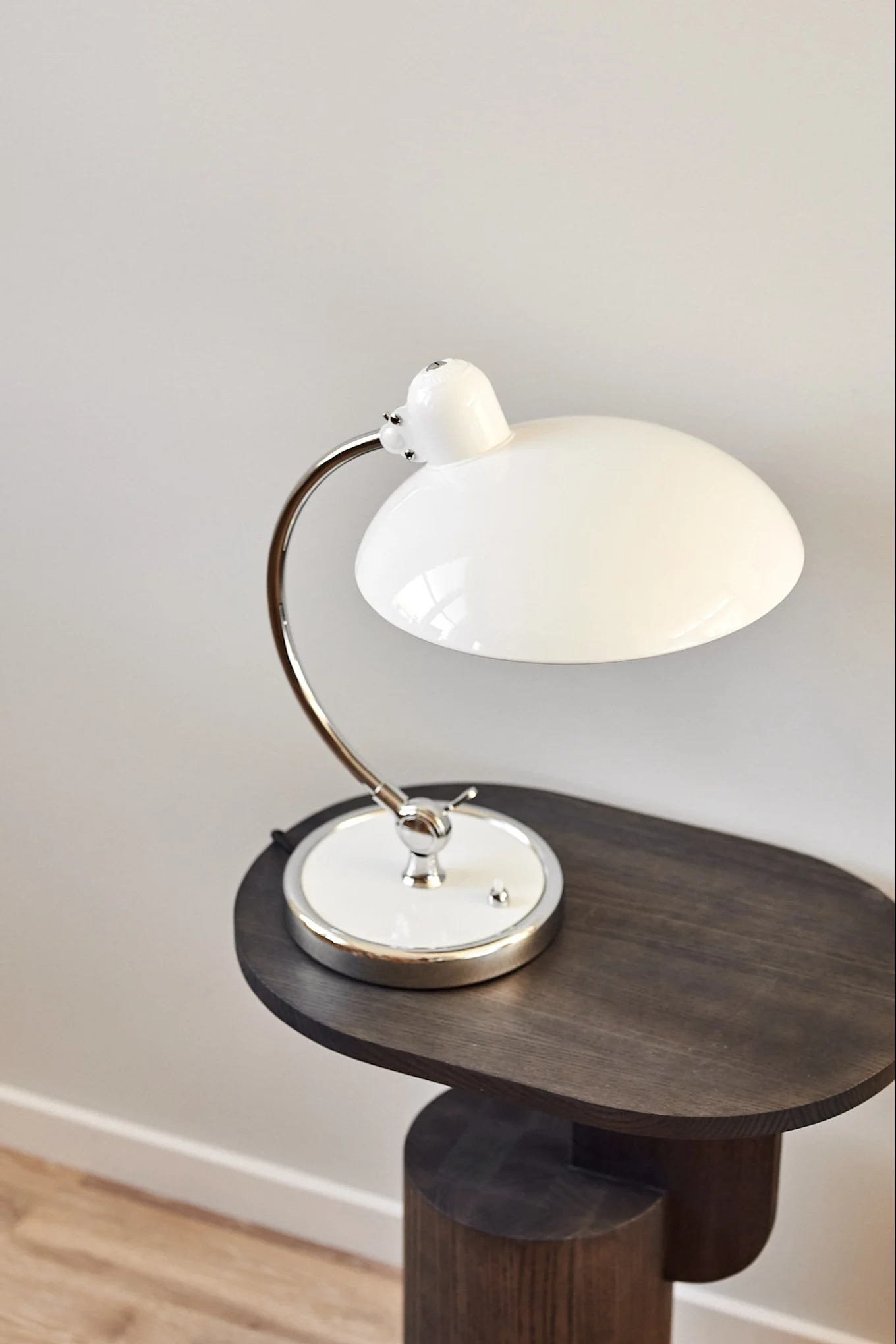 A white domed table lamp with a chrome curved arm sits on a dark, sculptural wooden side table.