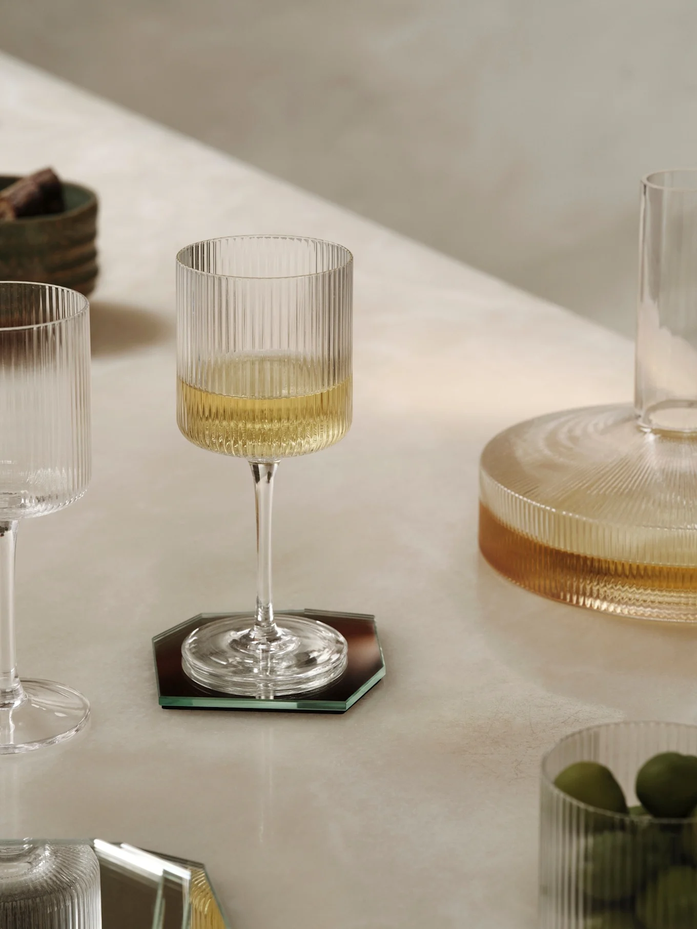A ribbed glass wine glass filled with white wine on a dark octagonal coaster, with other ribbed glass items and a ceramic bowl on a light table.