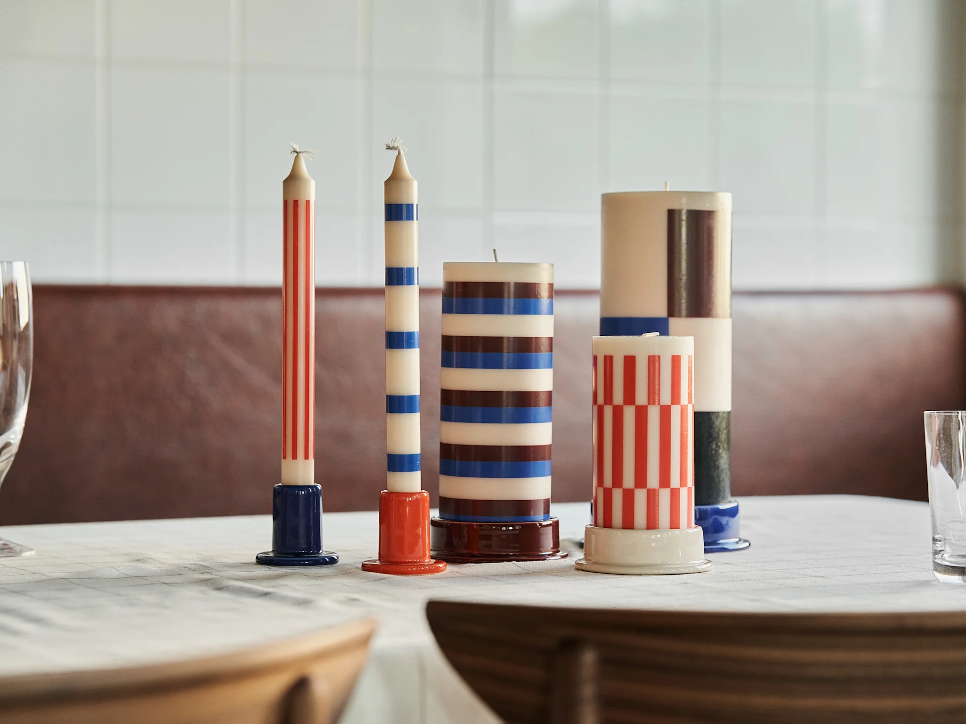 Five striped and checkered candles in red, blue, brown, and white on a dining table with ceramic holders.