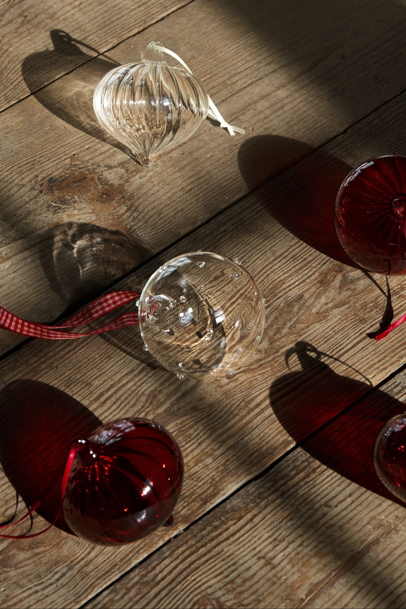 Clear and red glass Christmas ornaments with ribbons scattered on a sunlit wooden surface, creating shadows.