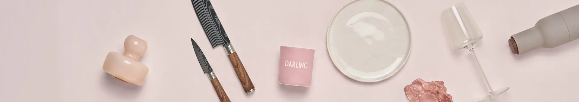 Assortment of kitchen and dining items on a pink background: a pink sculpture, two patterned knives, a 'DARLING' candle holder, a cream plate, a clear wine glass, and a grey bottle.