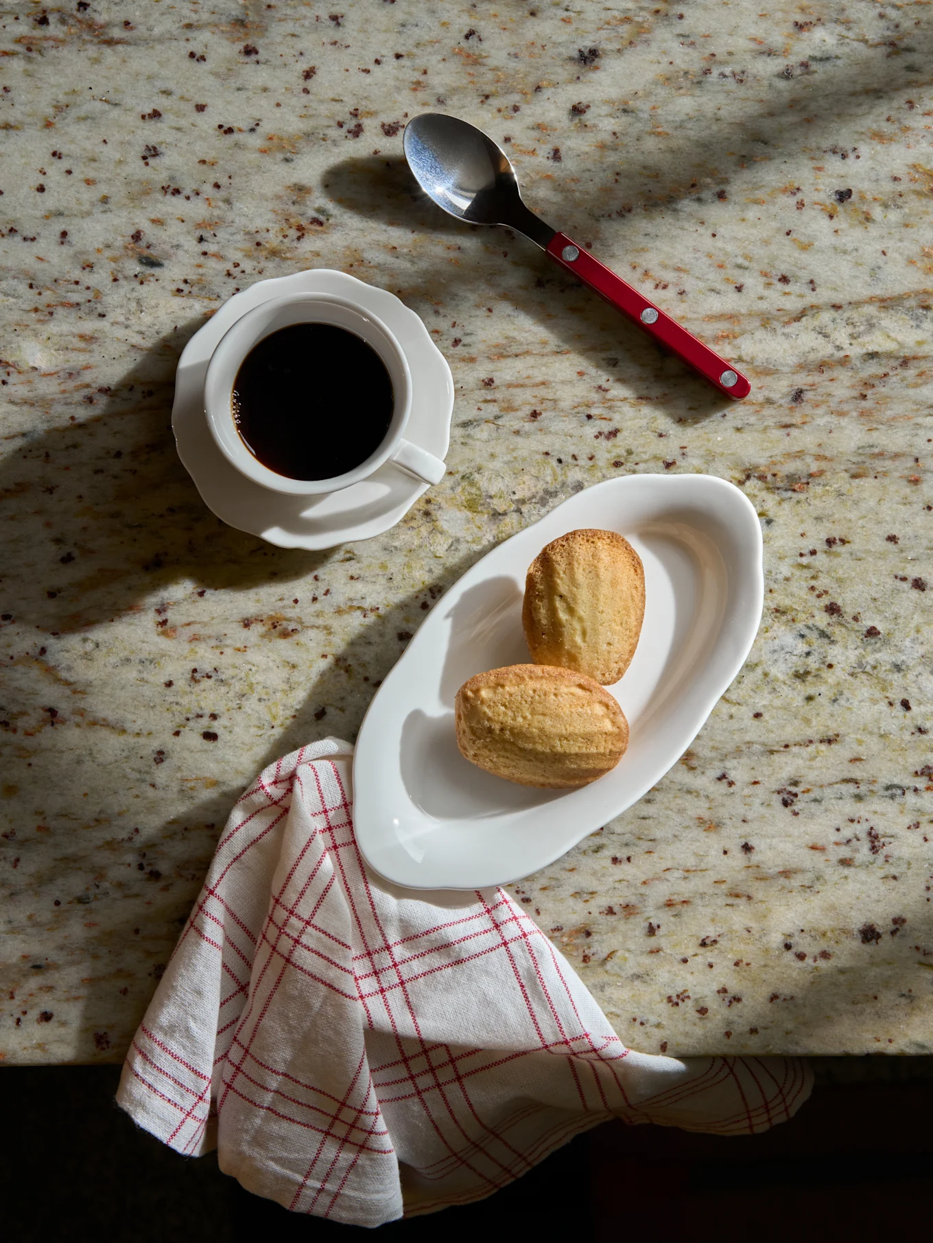 Overhead view of black coffee in a white cup, two madeleine cakes on a white plate, a spoon, and a striped napkin on a marble counter.