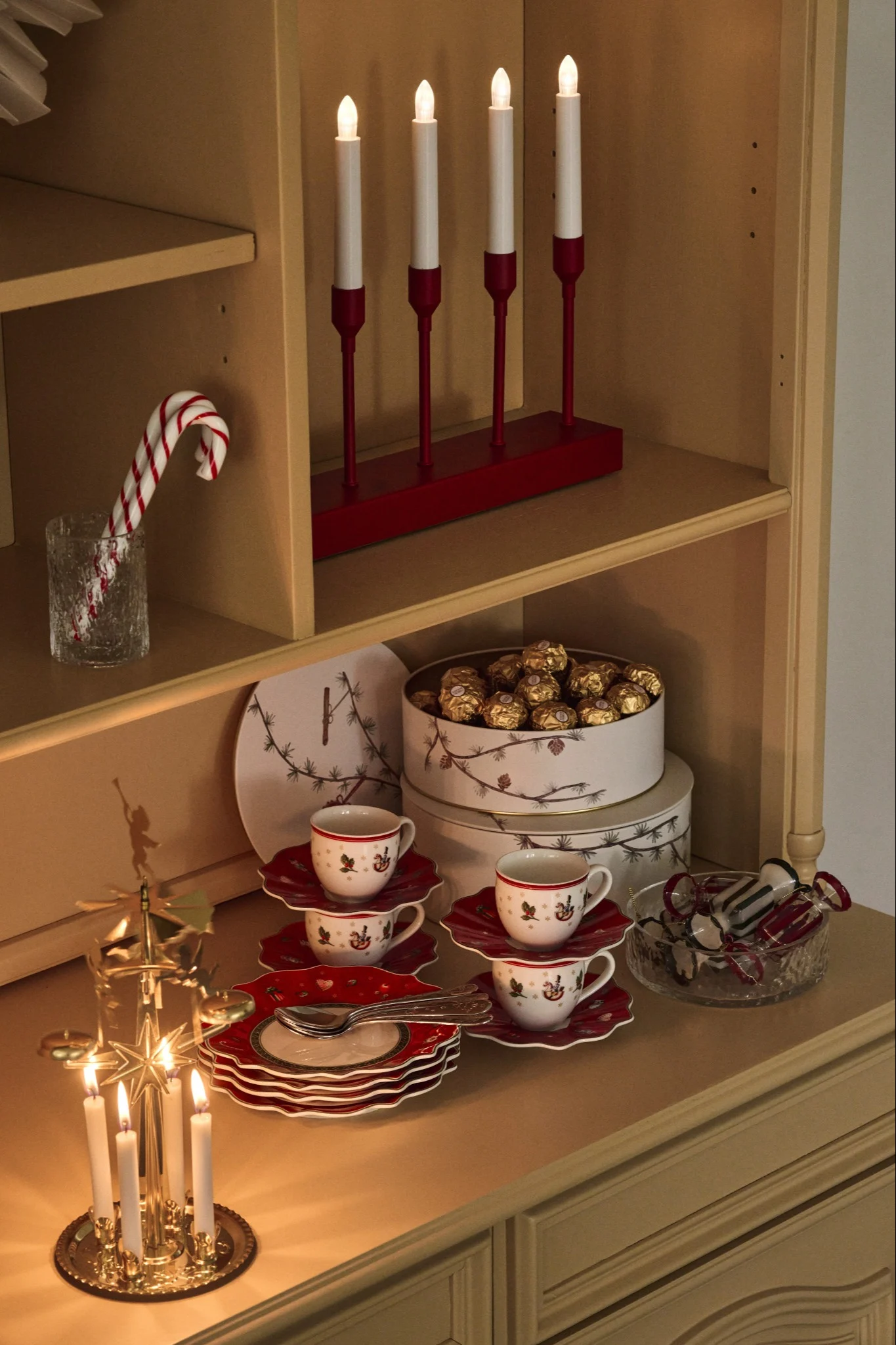 A hutch decorated for the holidays with lit candles, candy canes, chocolates, festive teacups, and decorative plates.