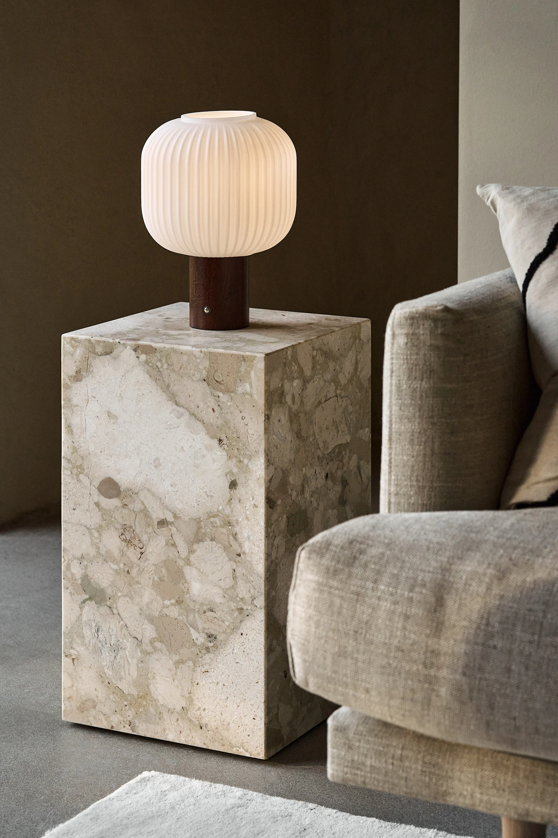 A modern white ribbed table lamp with a wooden base on a beige marble side table next to a sofa.