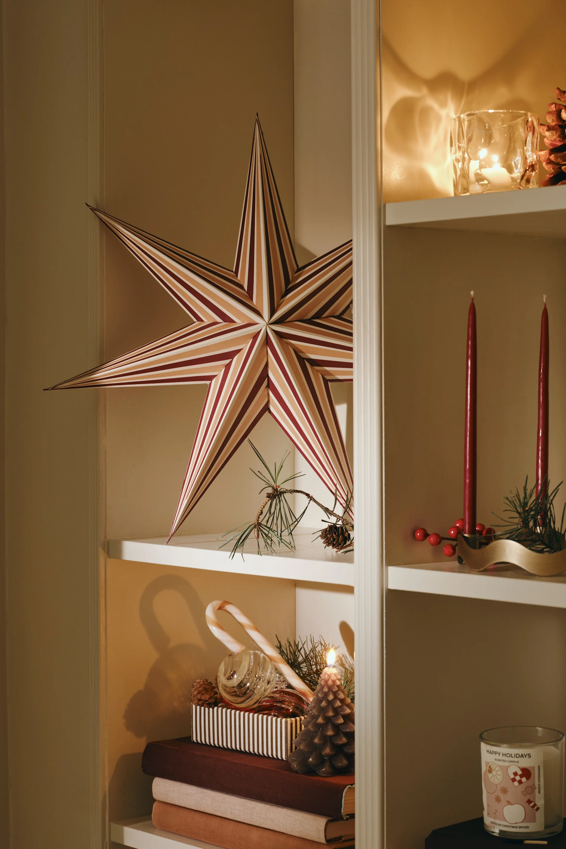 A white bookshelf adorned with holiday decor: a large striped star, red taper candles, a pinecone, a lit pinecone candle, a candy cane, and a "Happy Holidays" scented candle.