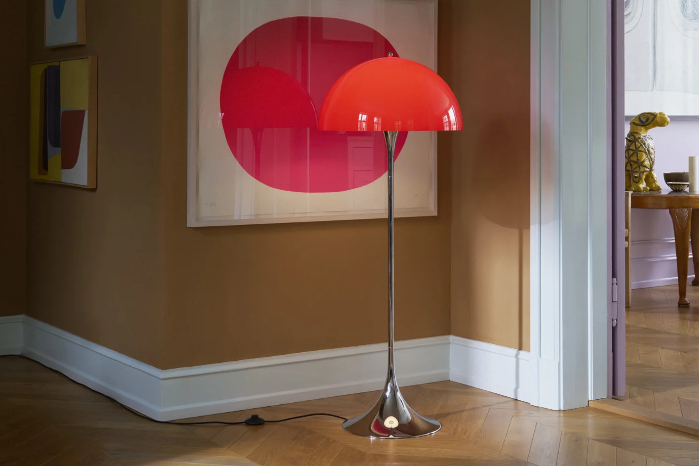 A bright red domed floor lamp with a chrome stand sits on a wooden floor in a room with brown walls and modern art.